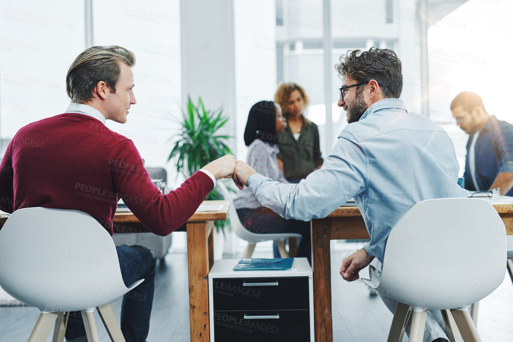 Buy stock photo Shot of two young colleagues giving each other a fist bump at their desks in a modern office