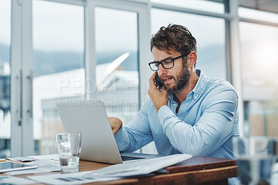 Buy stock photo Shot of a young businessman using a phone and laptop at his desk in a modern office