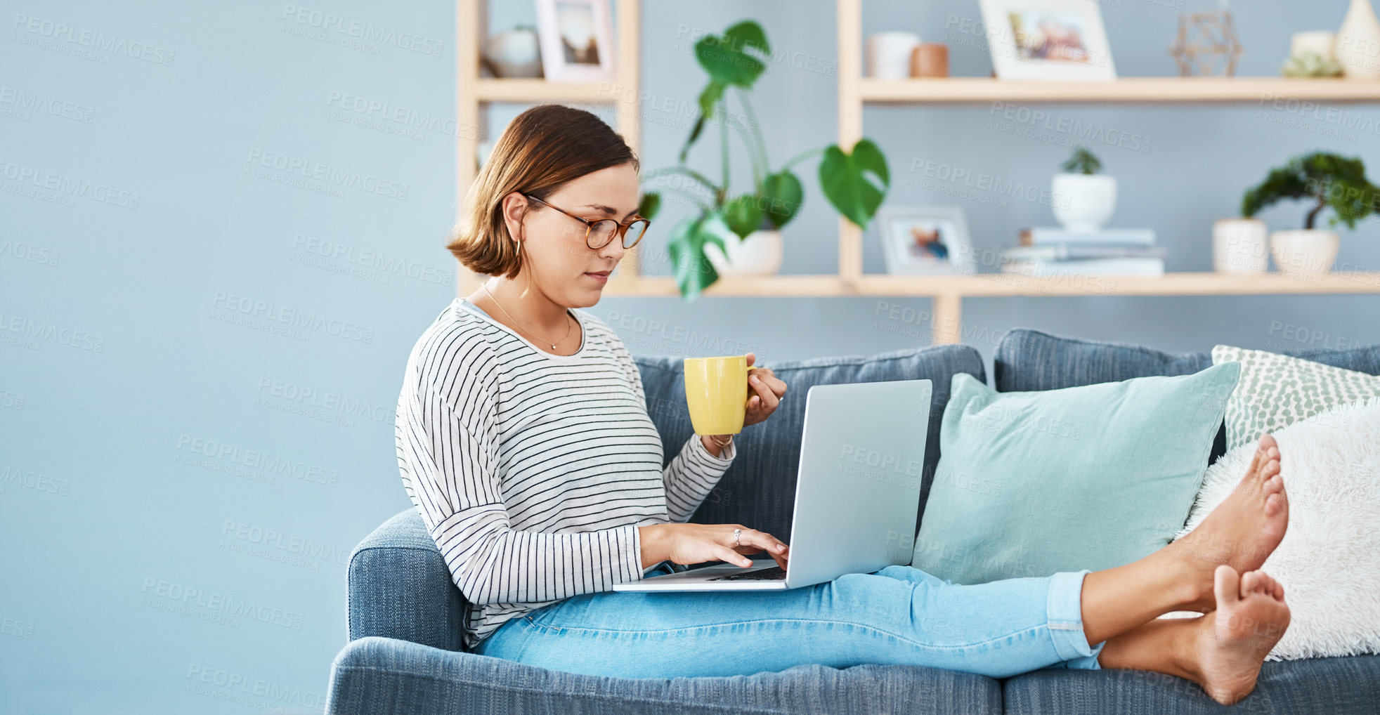 Buy stock photo Full length of a young woman using her laptop while having coffee in her living room