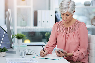 Buy stock photo Shot of a mature woman writing down details from her cellphone to her diary in her office at work