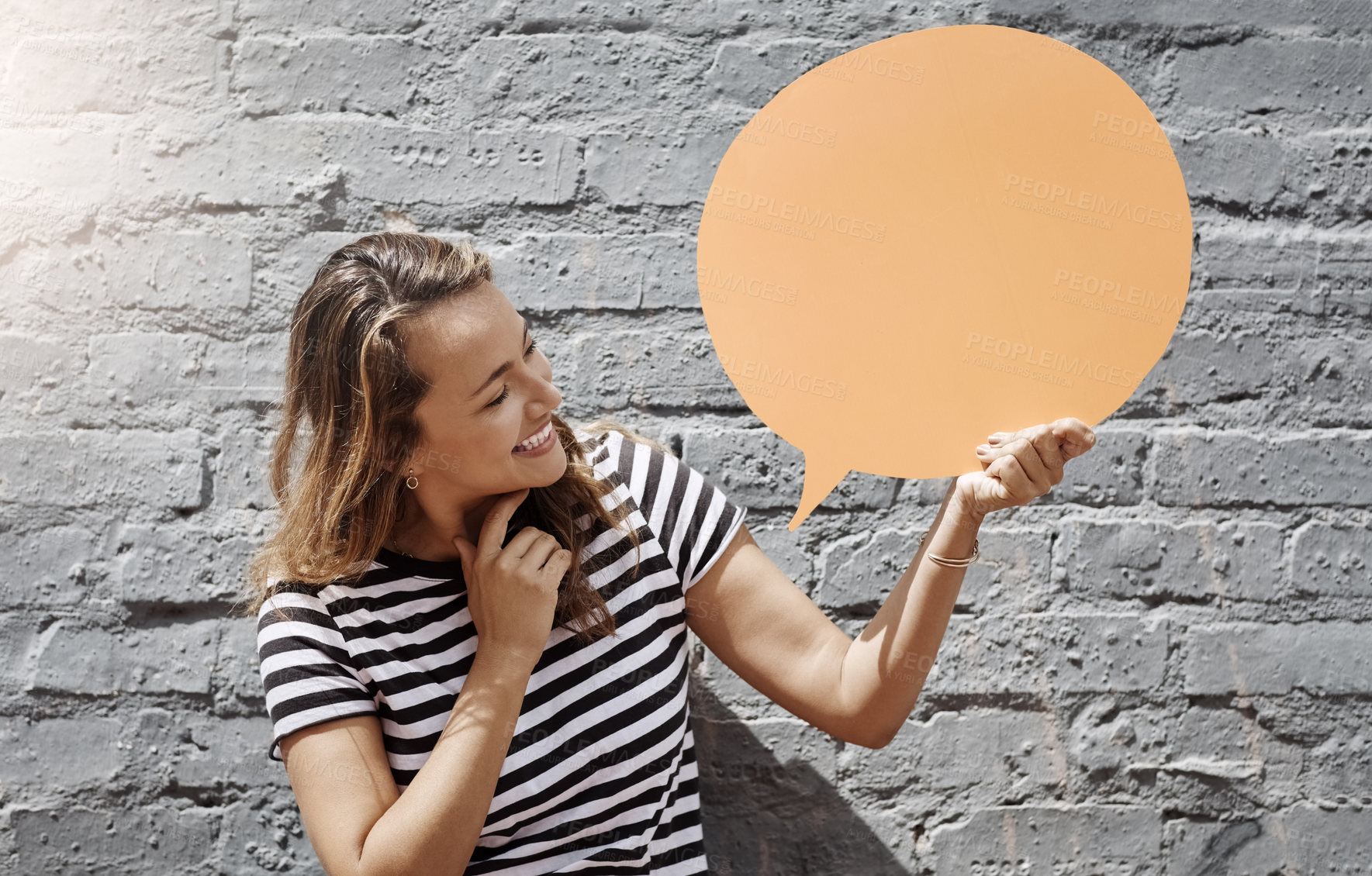 Buy stock photo Shot of a cheerful young woman holding a speech bubble against a brick wall outside