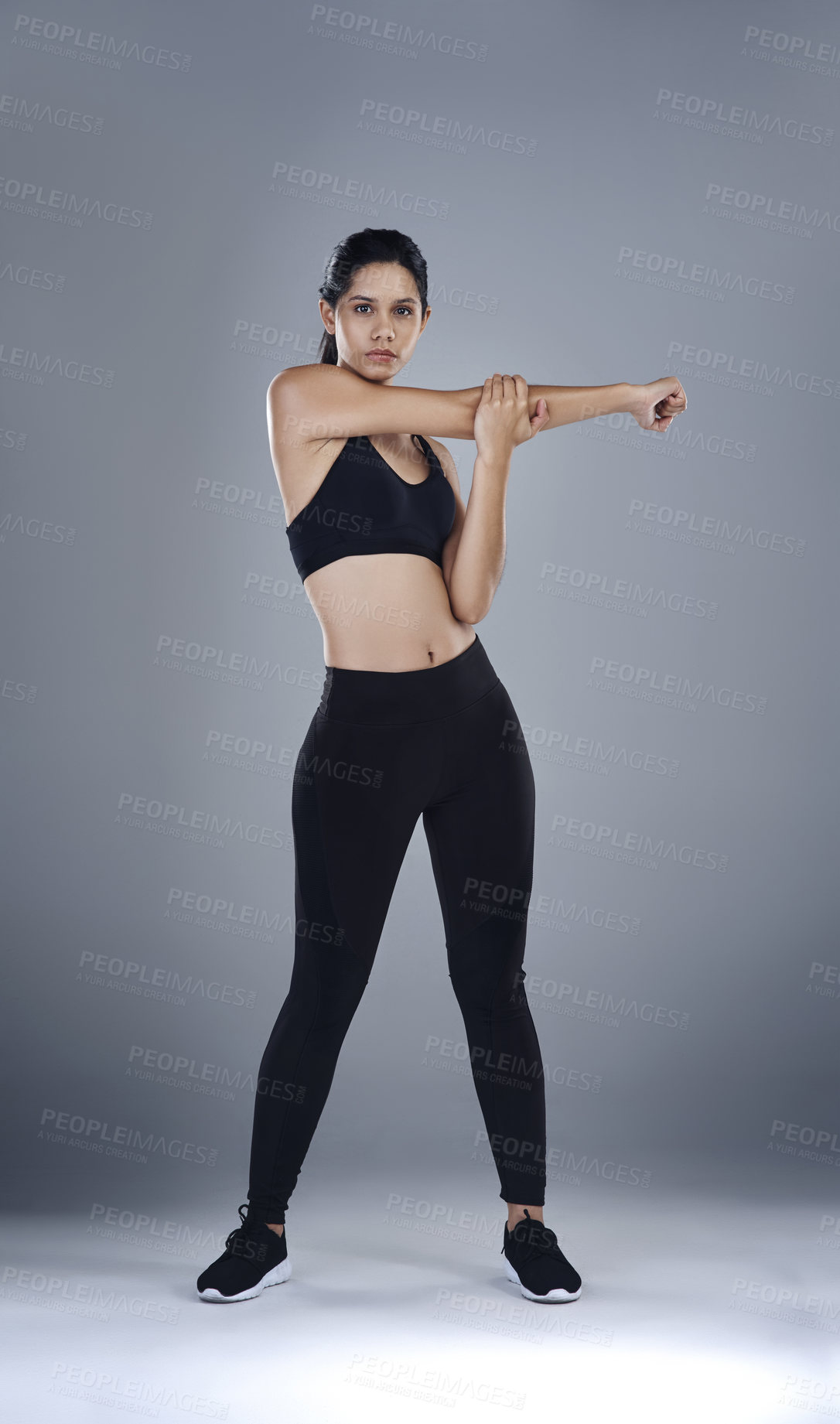 Buy stock photo Full length shot of a sporty young woman standing and stretching her arm against a grey background