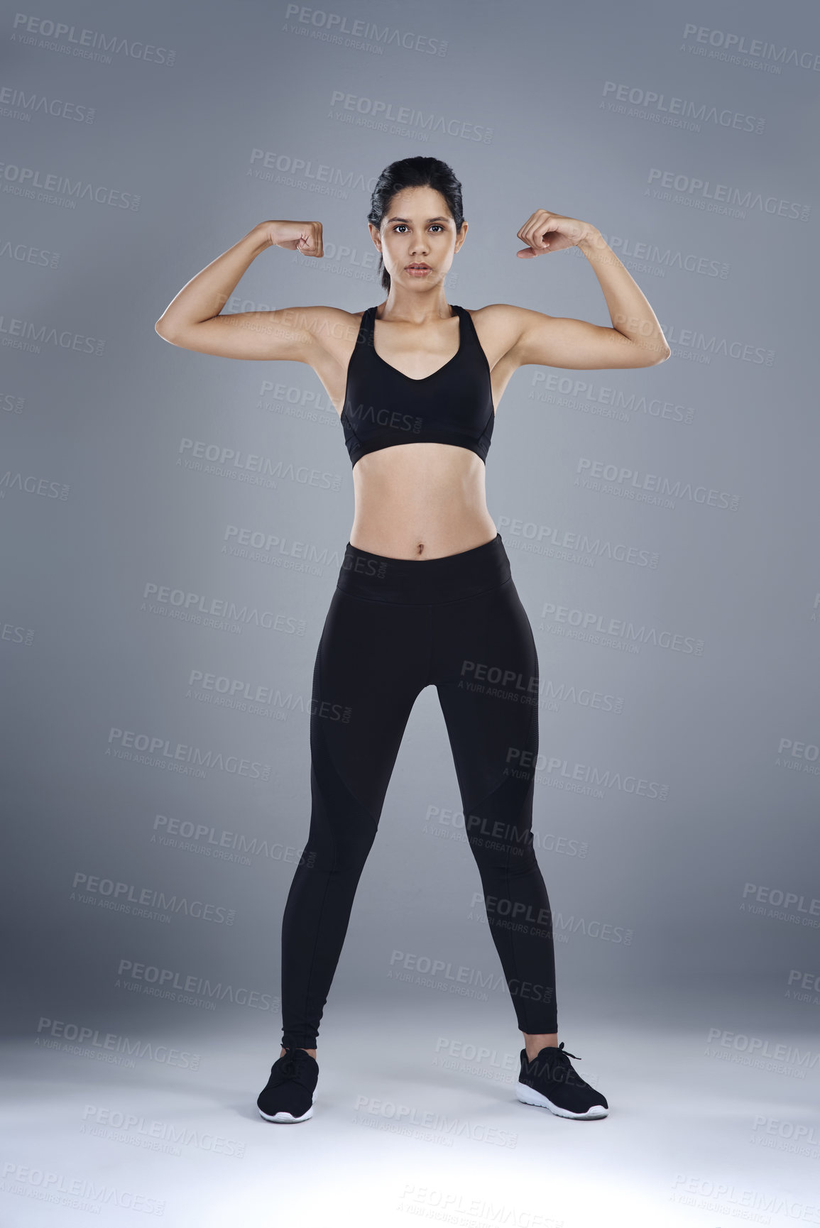 Buy stock photo Full length shot of a sporty young woman flexing her biceps against a grey background