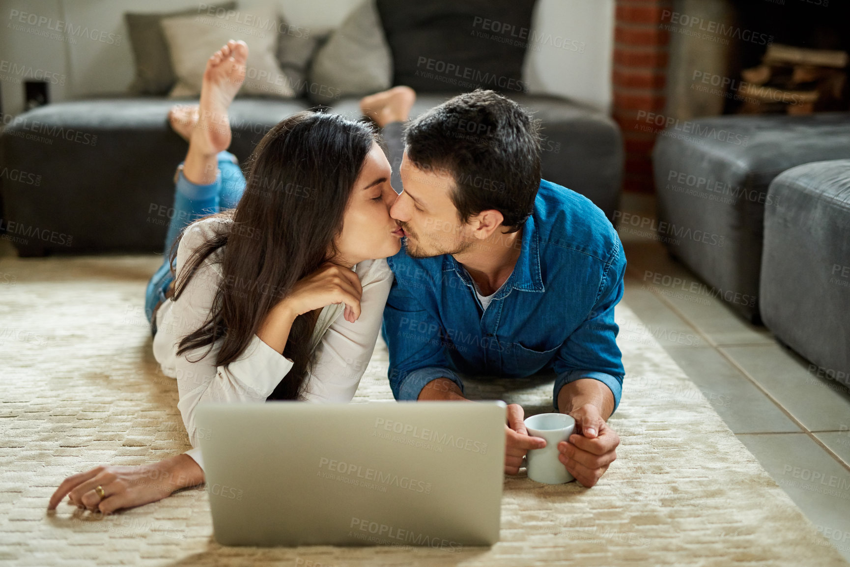 Buy stock photo Shot of an affectionate young couple kissing while lying down on the carpet and using their laptop at home