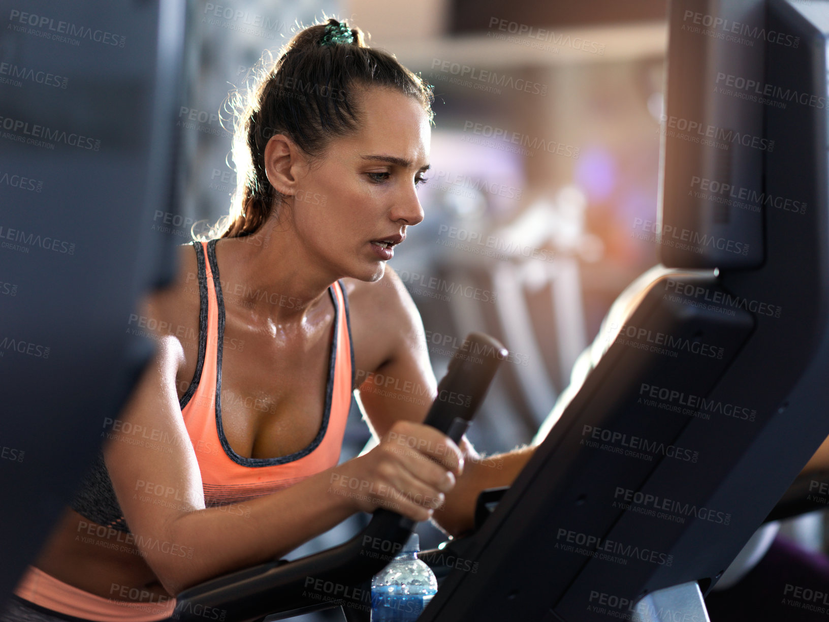 Buy stock photo Shot of a determined looking young woman working out on an elliptical machine in the gym