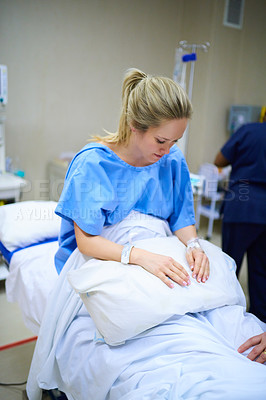 Buy stock photo Shot of a young pregnant woman sitting on her hospital bed and looking thoughtful with nurses in the background