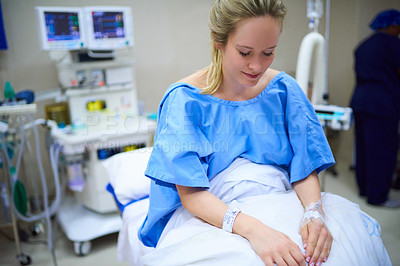 Buy stock photo Shot of a young pregnant woman sitting on her hospital bed and looking thoughtful with a nurse in the background