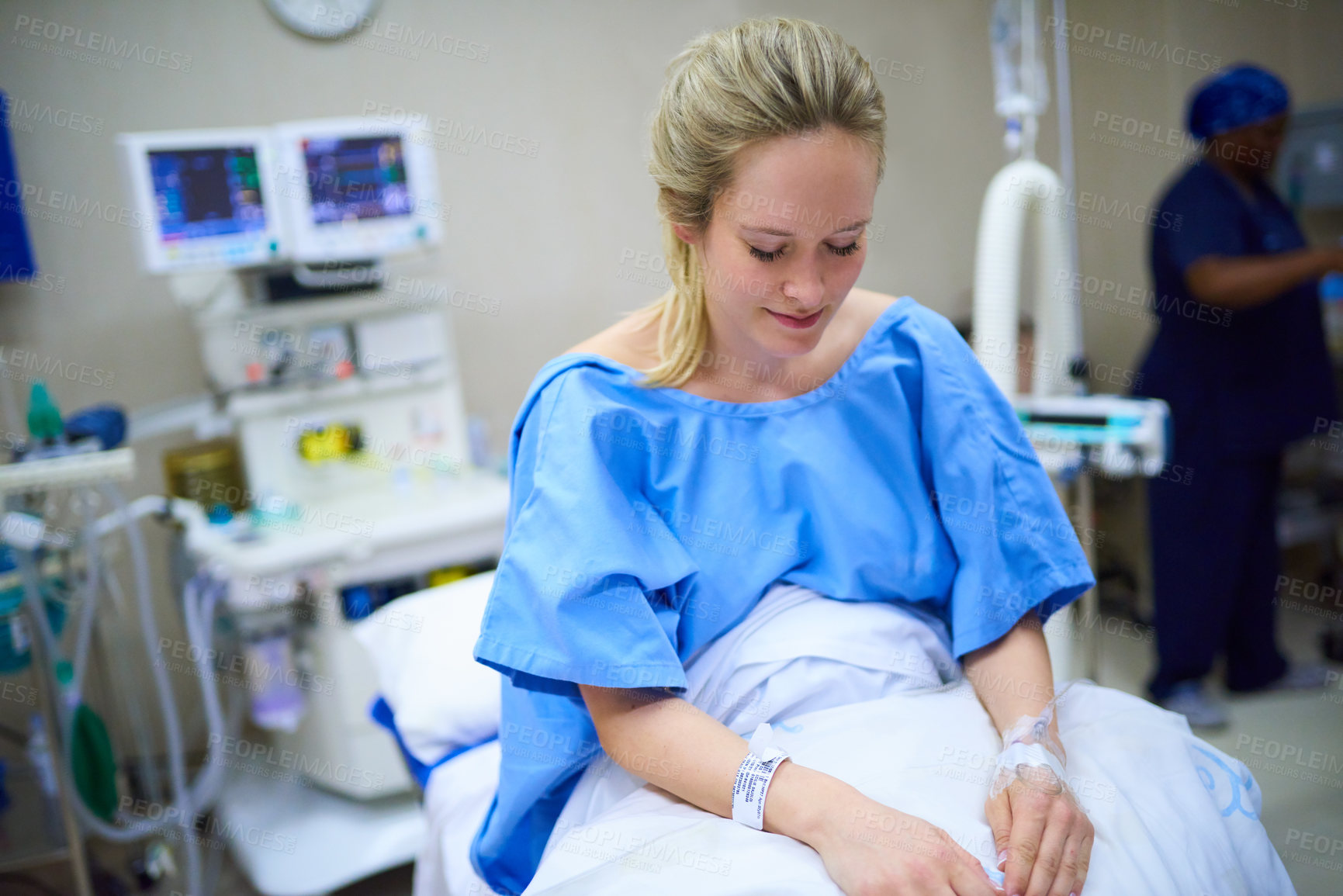 Buy stock photo Shot of a young pregnant woman sitting on her hospital bed and looking thoughtful with a nurse in the background