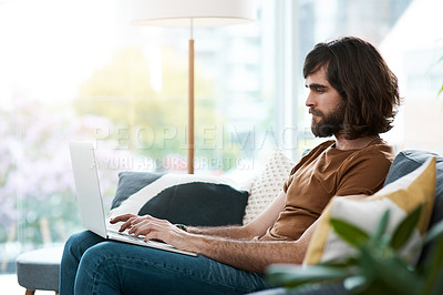 Buy stock photo Shot of a handsome young man using his laptop while relaxing on a sofa in his living room