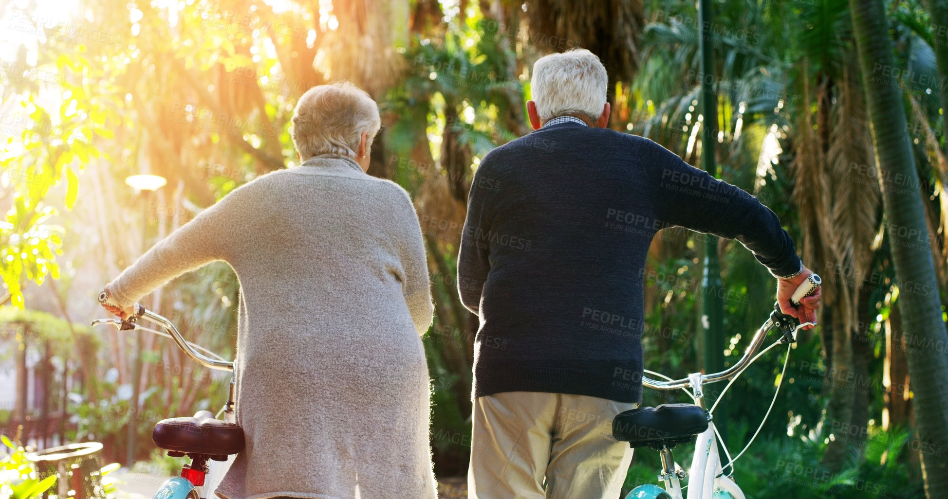 Buy stock photo Rearview shot of a elderly couple pushing their bicycles together outside in a park