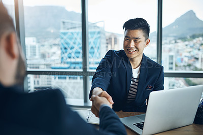 Buy stock photo Shot of two young businessmen shaking hands while sitting at a desk in a modern office