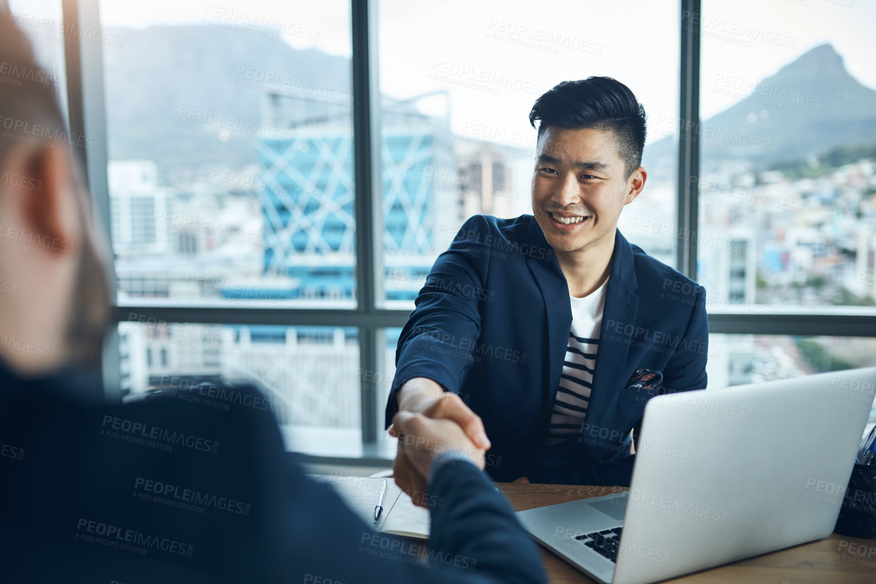 Buy stock photo Shot of two young businessmen shaking hands while sitting at a desk in a modern office