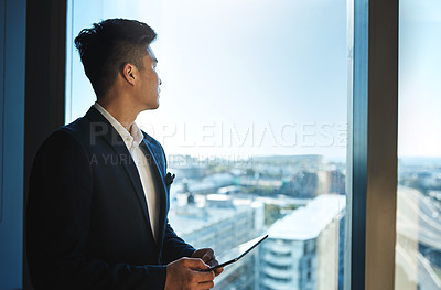 Buy stock photo Shot of a young businessman using a digital tablet while looking out the window in an office