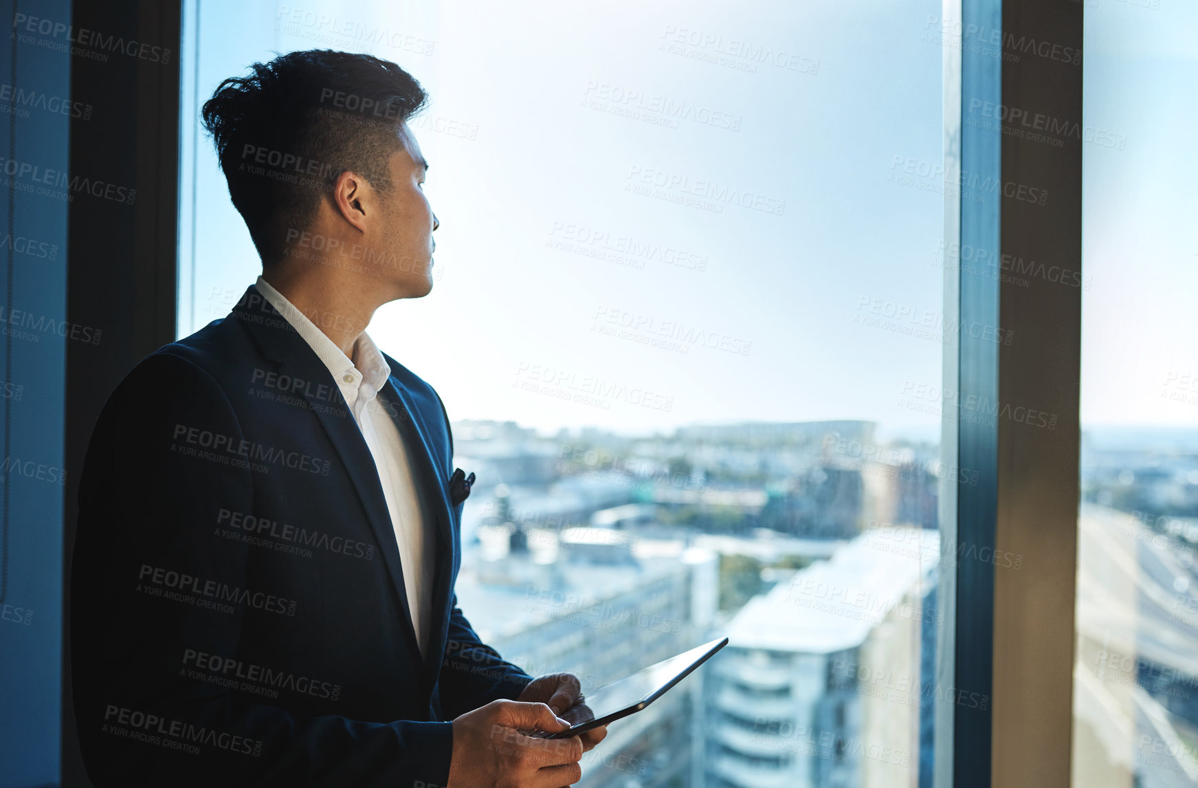 Buy stock photo Shot of a young businessman using a digital tablet while looking out the window in an office