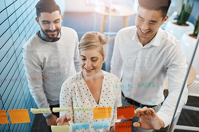 Buy stock photo Cropped shot of three young businesspeople brainstorming with sticky notes on a glass wall in the office