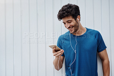 Buy stock photo Shot of a sporty young man standing against a white wall after exercising and listening to music outside during the day
