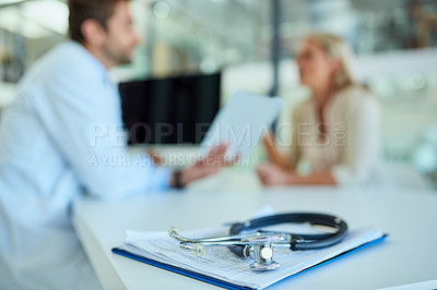 Buy stock photo Shot of a stethoscope and a clipboard on a desk with a doctor and patient in the background
