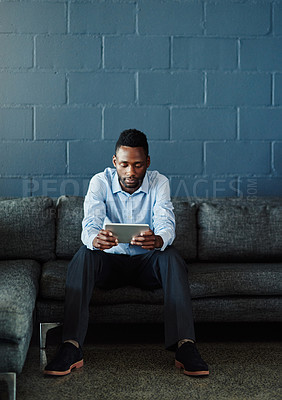 Buy stock photo Full length shot of a handsome young businessman using his tablet while sitting on a sofa in the office