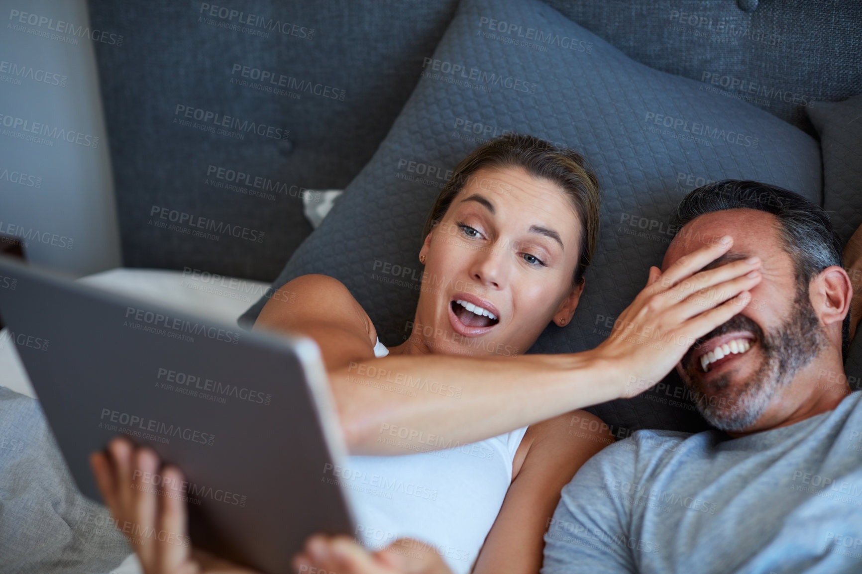 Buy stock photo High angle shot of an attractive young woman covering her husband's eyes while holding a digital tablet