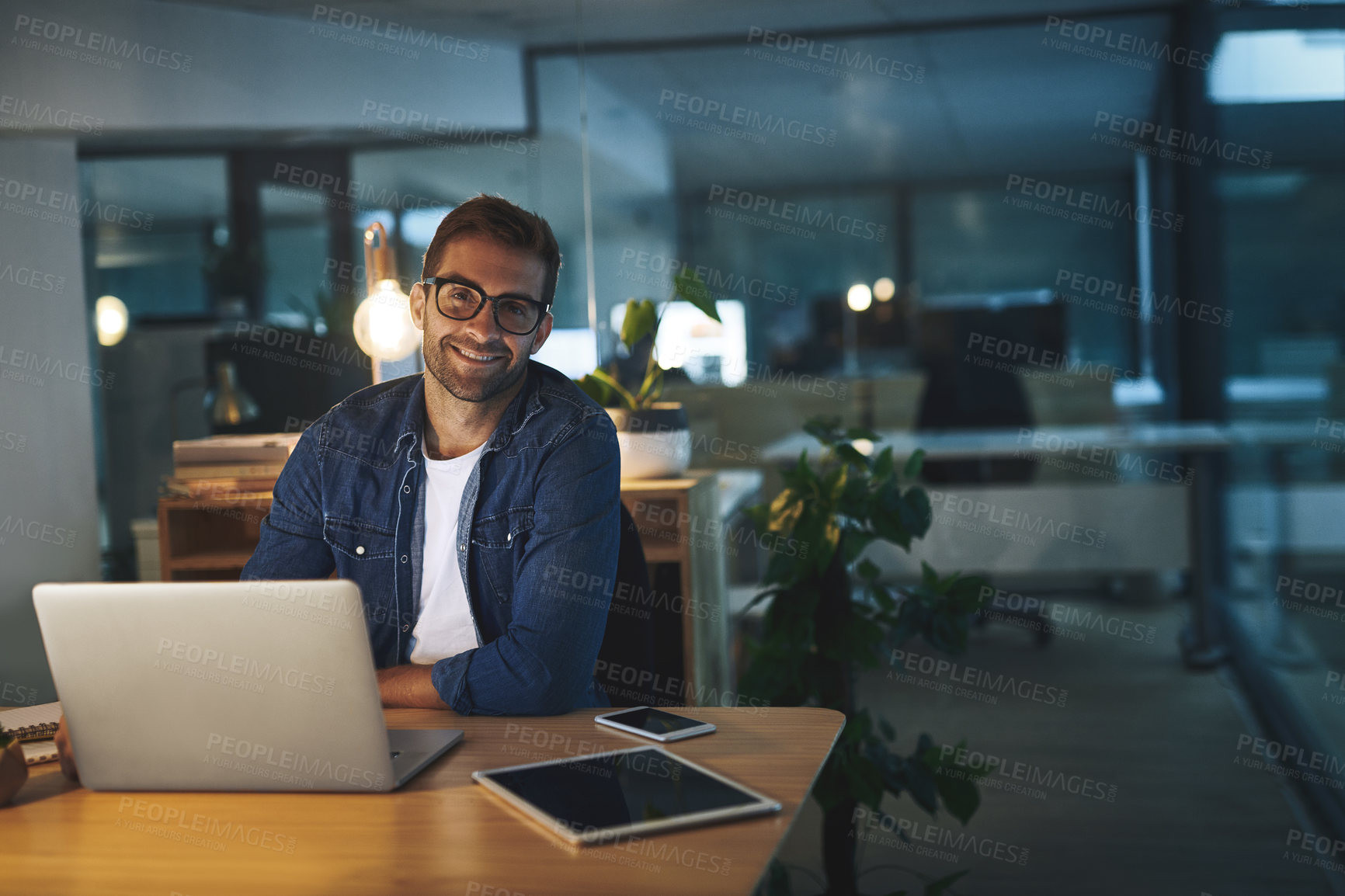 Buy stock photo Portrait of a handsome young businessman smiling while working late at night in a modern office