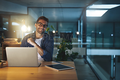 Buy stock photo Portrait of a handsome young businessman smiling while working late at night on his computer in a modern office