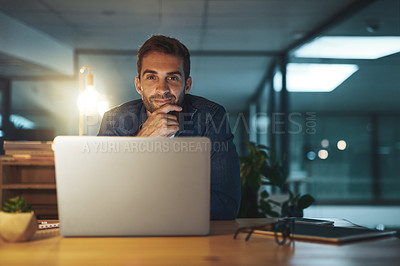 Buy stock photo Portrait of a handsome young businessman smiling while working late at night on his computer in a modern office