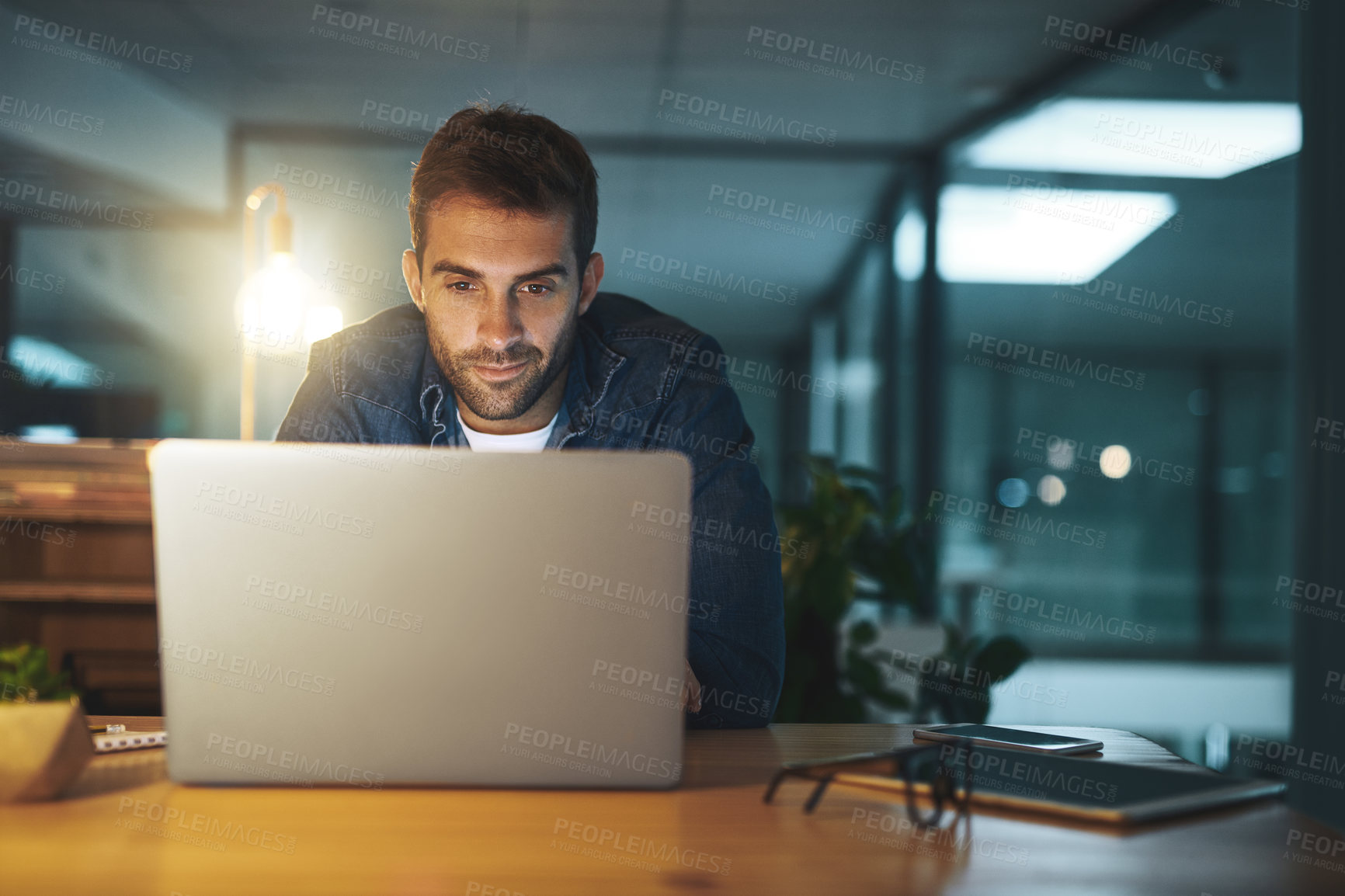 Buy stock photo Shot of a handsome young businessman working late at night on his computer in a modern office