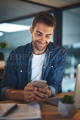 Buy stock photo Shot of a young handsome businessman smiling while using his cellphone at his desk in a modern office