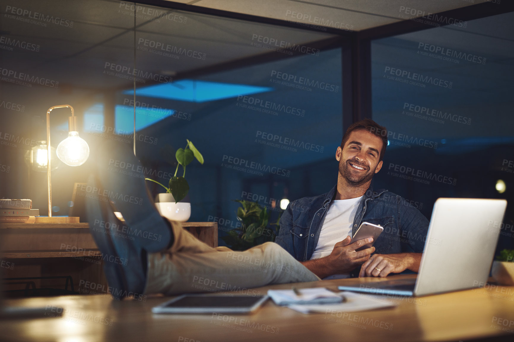 Buy stock photo Night, office and portrait of happy man with phone, laptop and confidence at desk at digital agency. Late, relax and creative consultant with smartphone for scroll, communication or business contact