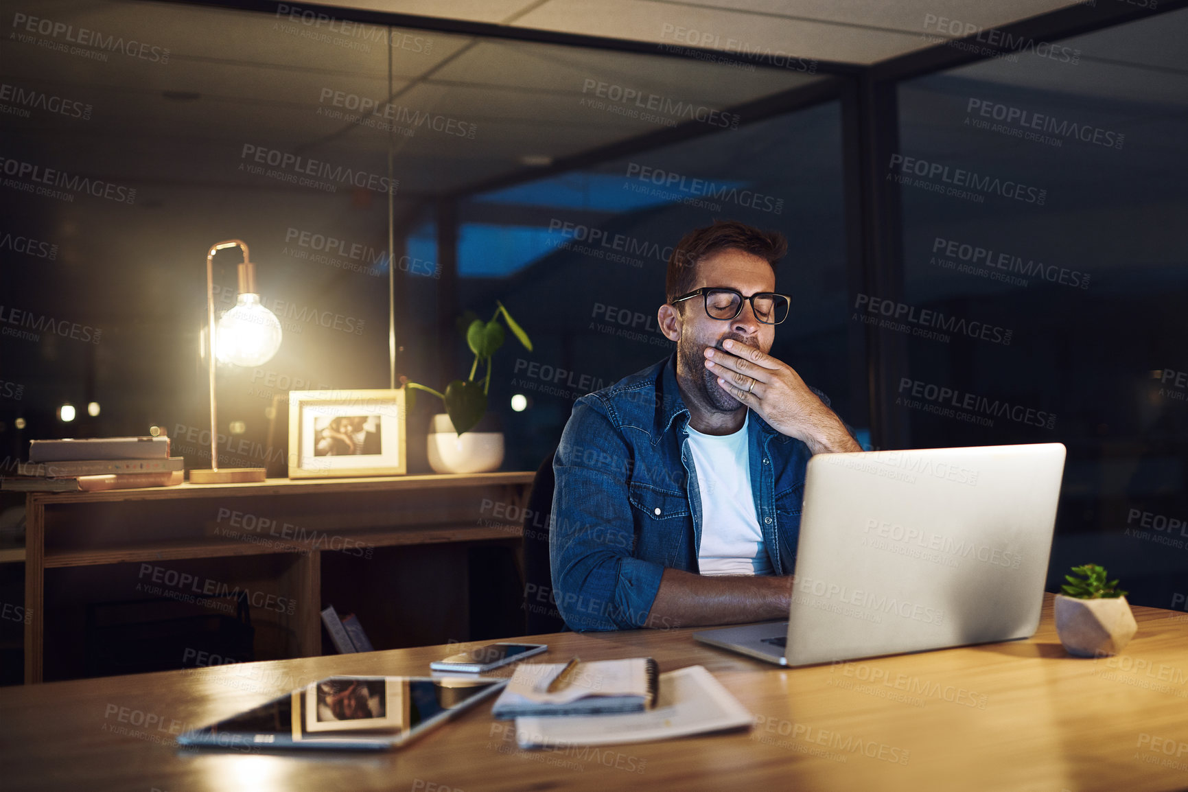 Buy stock photo Shot of a handsome young businessman yawning while working late at night in a modern office