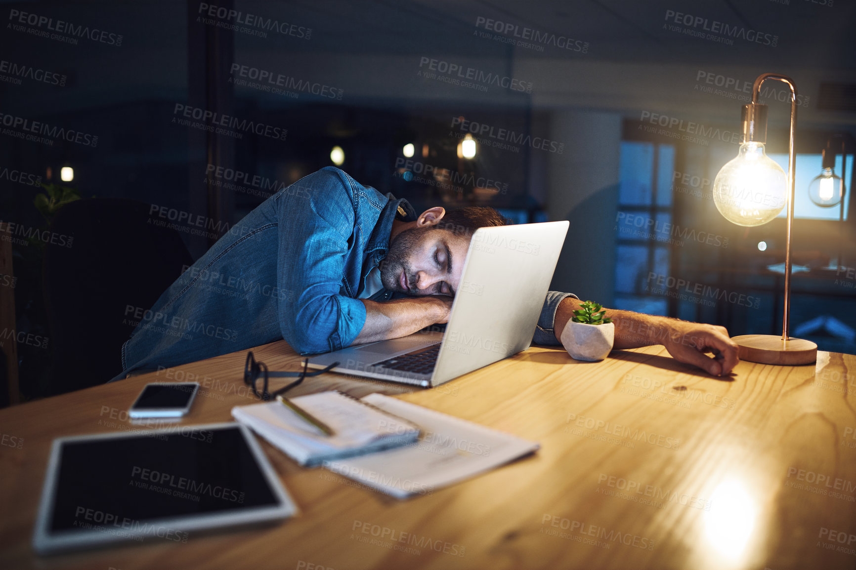 Buy stock photo Shot of a handsome young businessman asleep at his desk while working late at night in a modern office