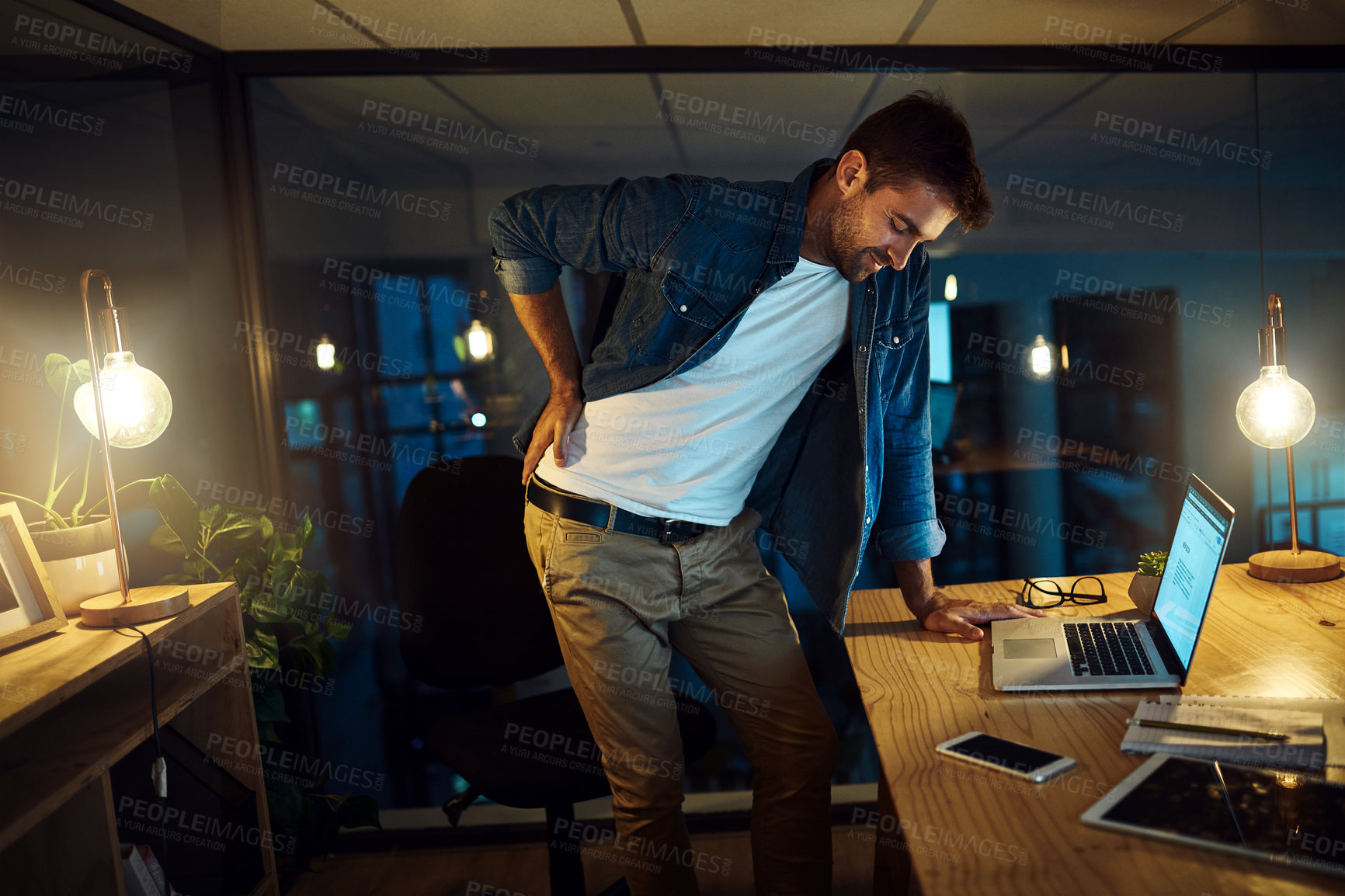 Buy stock photo Shot of a handsome young businessman standing up to stretch his back while working late at night in a modern office