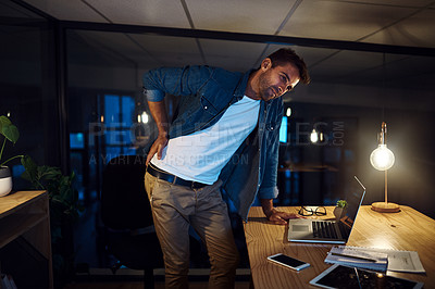 Buy stock photo Shot of a handsome young businessman standing up to stretch his back while working late at night in a modern office