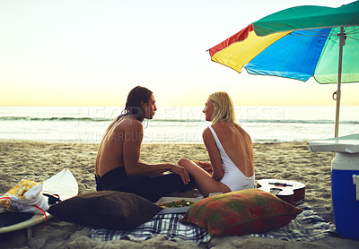 Buy stock photo Rearview shot of an affectionate young couple having a picnic date on the beach at sunset