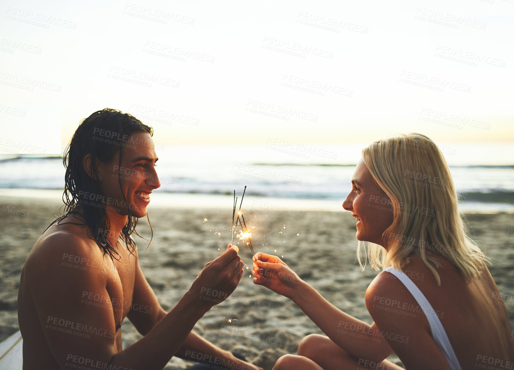 Buy stock photo Shot of a young happy couple having fun and lighting sparklers while on a date on the beach at sunset