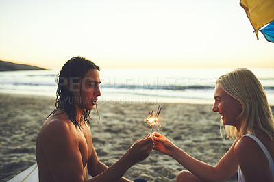 Buy stock photo Shot of a happy young couple having fun and lighting sparklers while on a date on the beach at sunset
