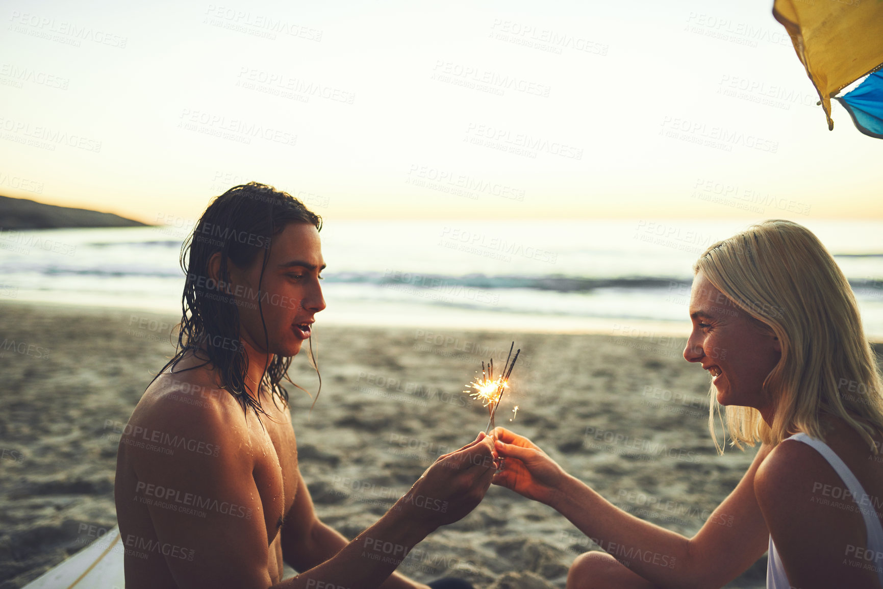 Buy stock photo Shot of a happy young couple having fun and lighting sparklers while on a date on the beach at sunset