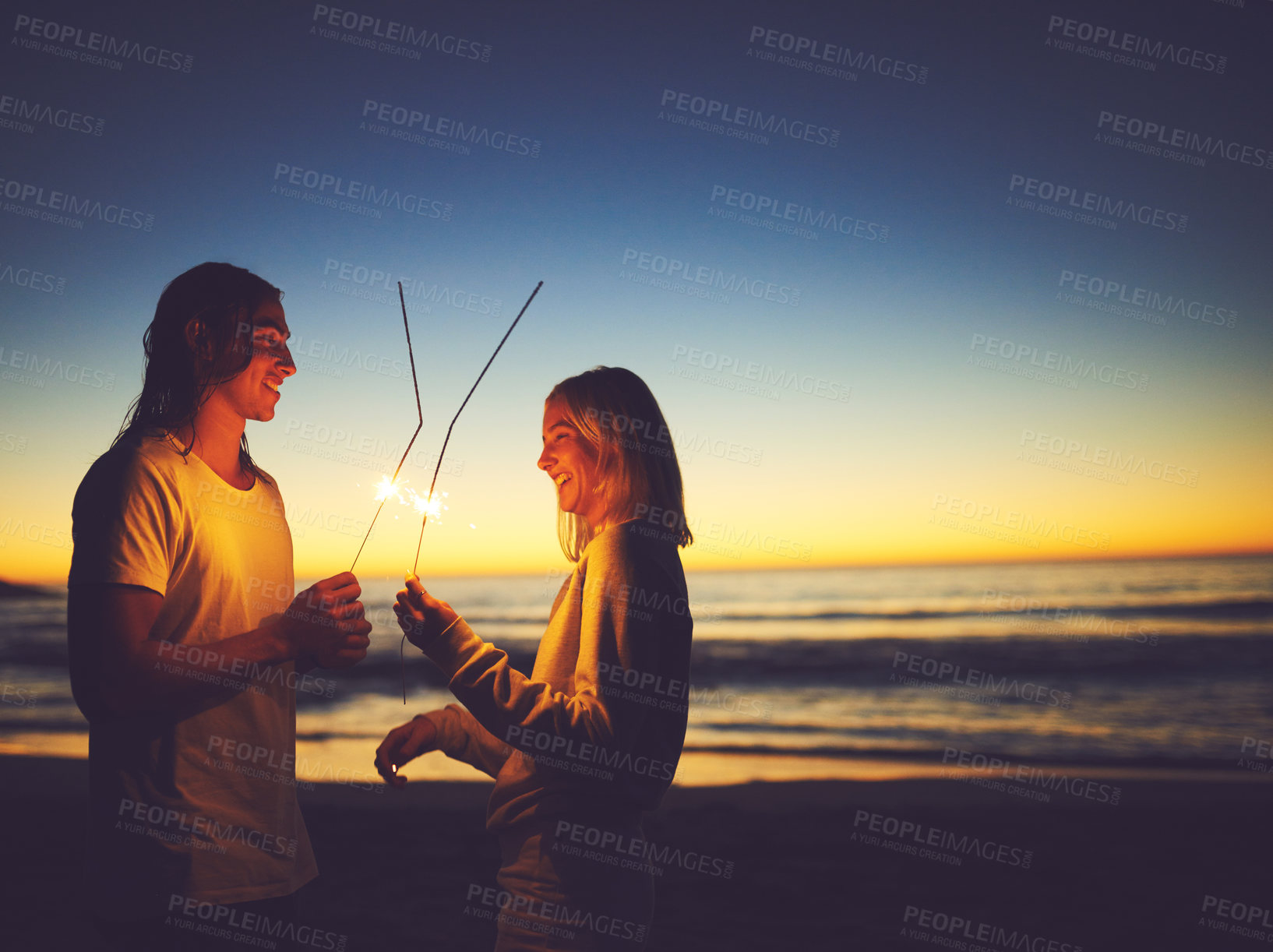 Buy stock photo Shot of a young couple playing with sparklers on the beach at night