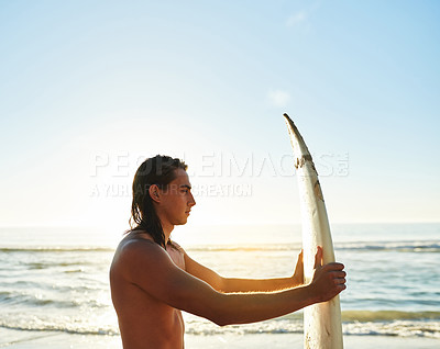 Buy stock photo Cropped shot of a handsome young man holding and looking at his surfboard on the beach at sunset