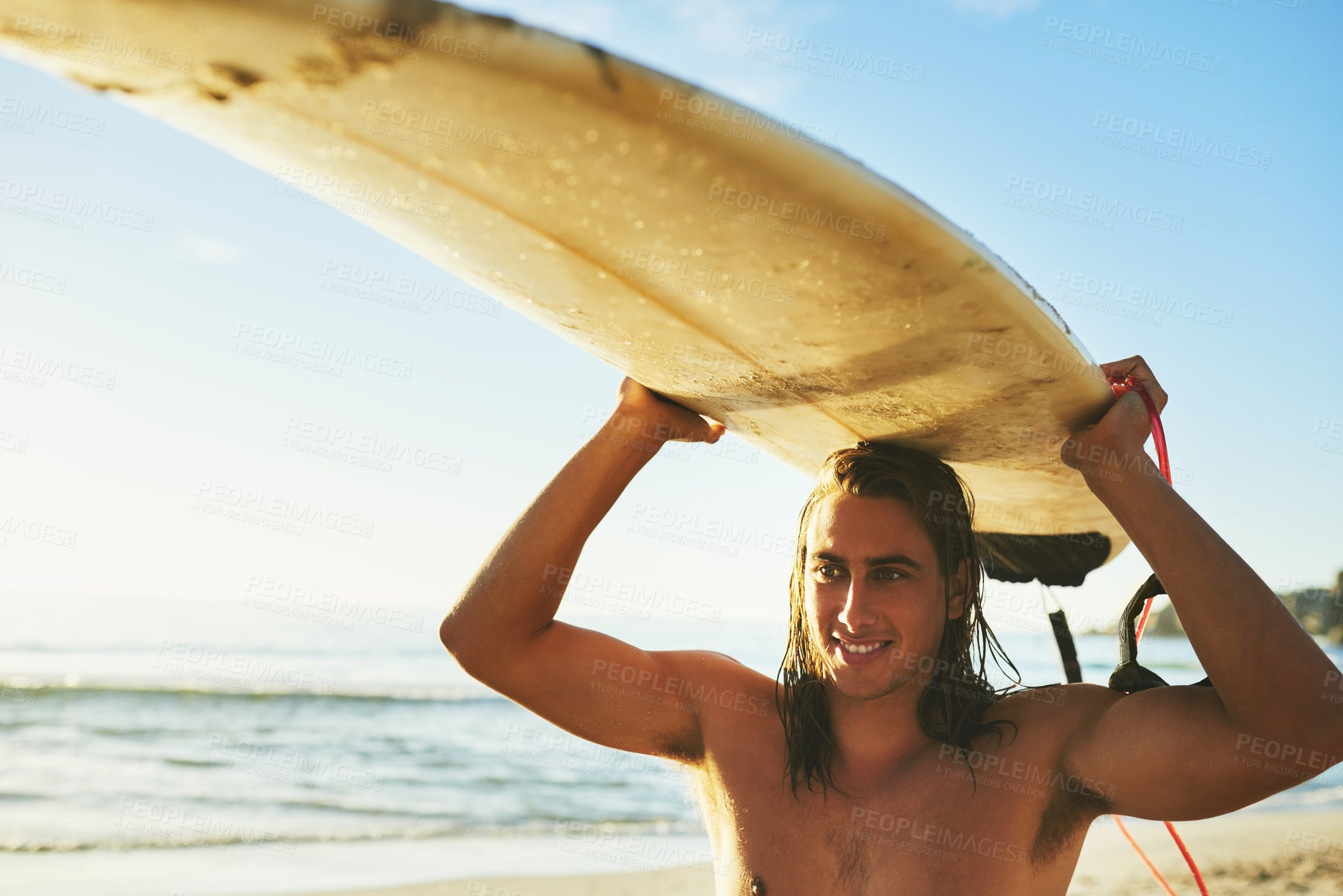 Buy stock photo Cropped shot of a handsome young man smiling while carrying his surfboard over his head on the beach at sunset