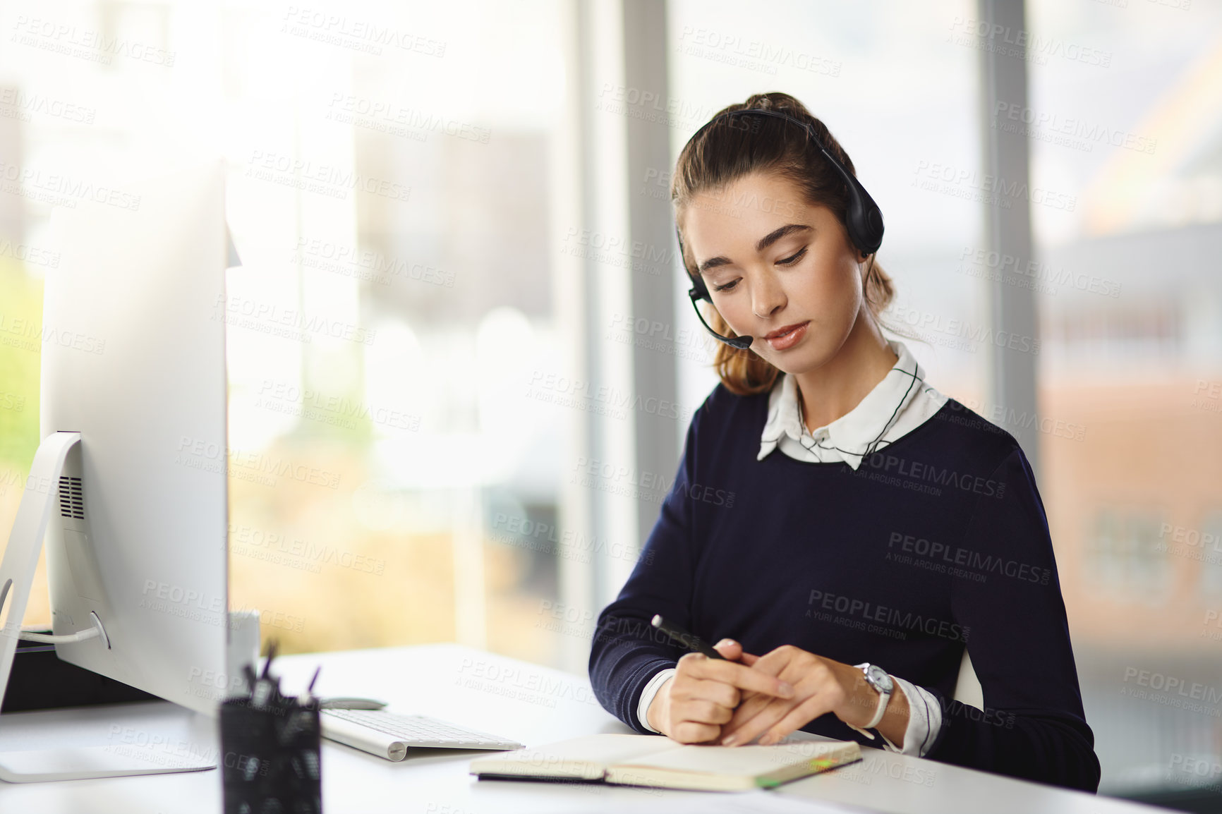 Buy stock photo Shot of an attractive young businesswoman wearing a headset and writing notes at her desk in a modern office