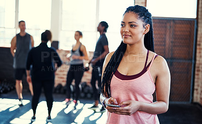 Buy stock photo Shot of a cheerful young woman standing and using her cellphone while contemplating before a workout in a gym