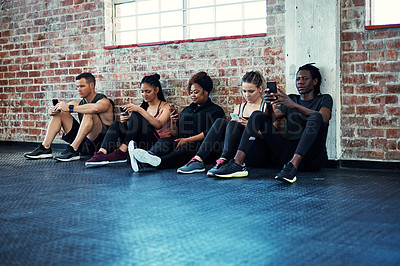 Buy stock photo Shot of a cheerful young group of people sitting down the floor and using their cellphones before a workout in a gym