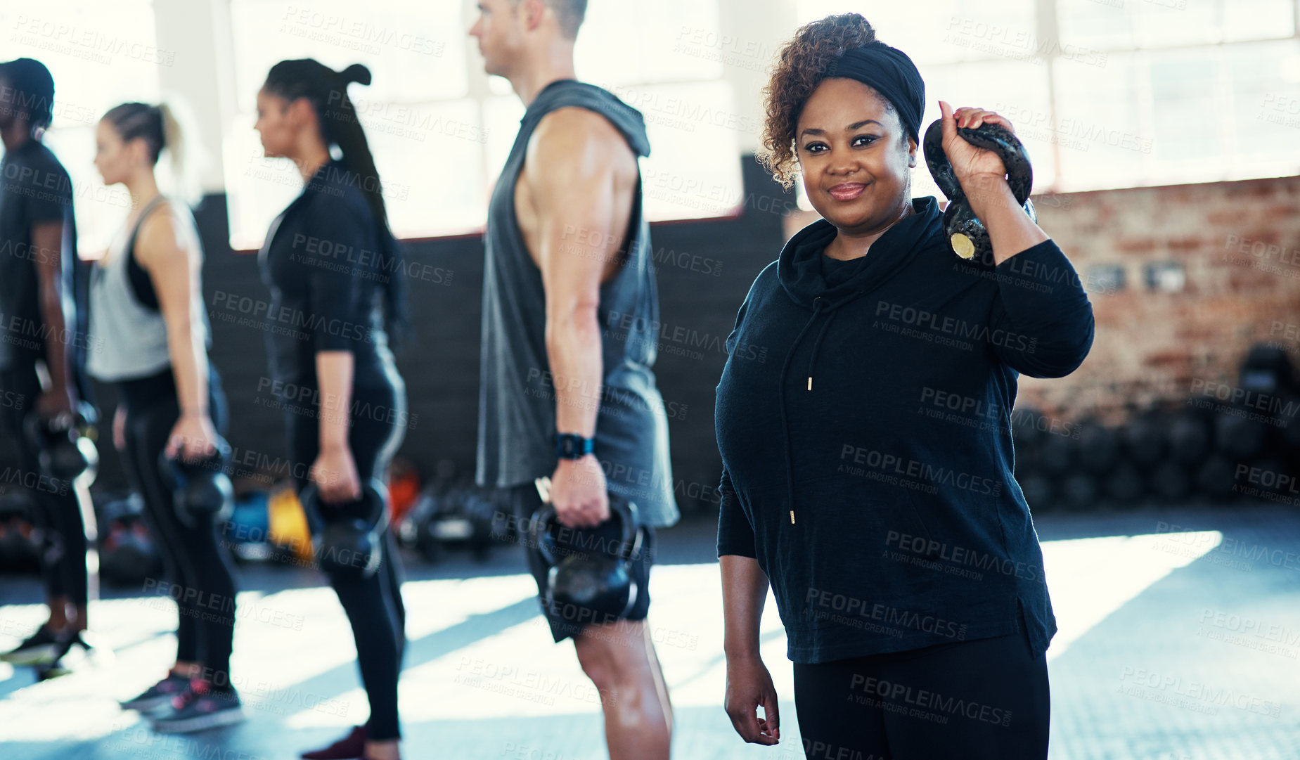 Buy stock photo Shot of a cheerful group of young people standing in a row and training with weights  while one looks into the camera in a gym