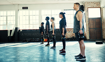 Buy stock photo Shot of a focused group of young people standing in a row and training with weights in a gym