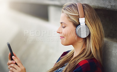 Buy stock photo Shot of an attractive young woman leaning against a wall and listening to music while using her cellphone