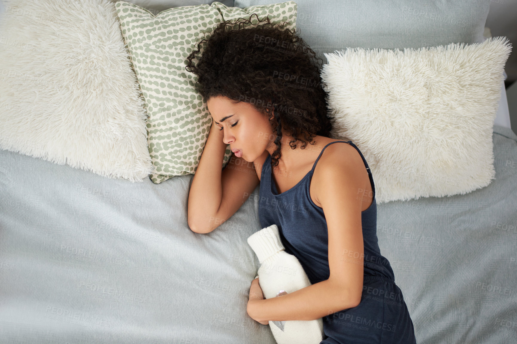 Buy stock photo Bed, hot water bottle and black woman with cramps, period and pain of stomach ache, sick and morning in house. Home, abdomen and person in bedroom, sleeping and girl with discomfort of menstruation