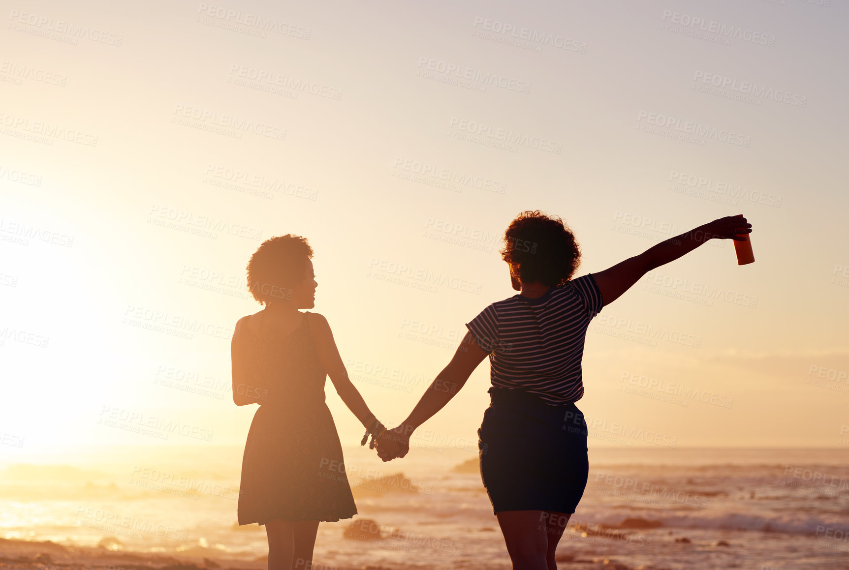 Buy stock photo Rearview shot of two unrecognizable young women holding hands and walking along the beach at sunset