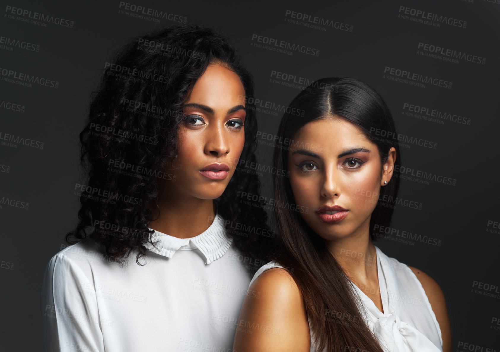 Buy stock photo Portrait of two attractive young women wearing white blouses posing closely together against a dark background in the studio