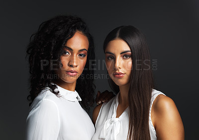 Buy stock photo Portrait of two attractive young women wearing white blouses posing closely together against a dark background in the studio